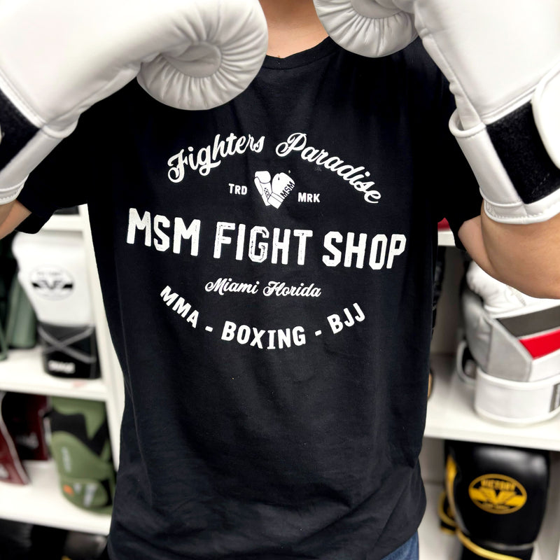 Person wearing a black t-shirt with 'MSM Fight Shop' text, white boxing gloves, and gym equipment in the background.