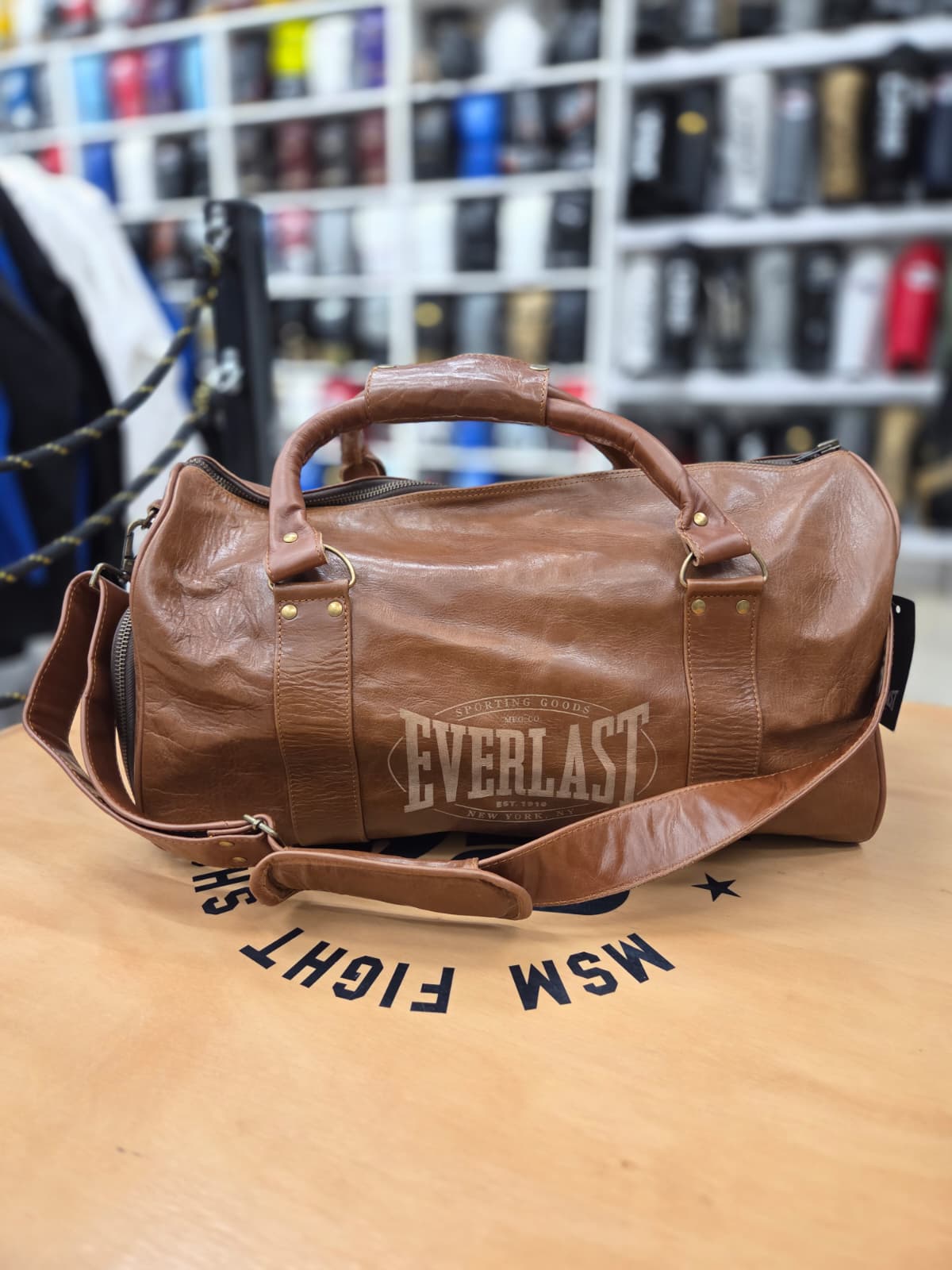 A brown leather Everlast duffel bag with a top handle and shoulder strap, resting on a wooden table in a retail store setting.