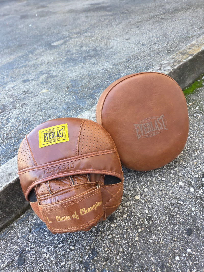 A pair of brown Everlast leather boxing gloves and a matching round protective pad resting on an asphalt surface. The Everlast brand is clearly visible on the gloves and pad.
