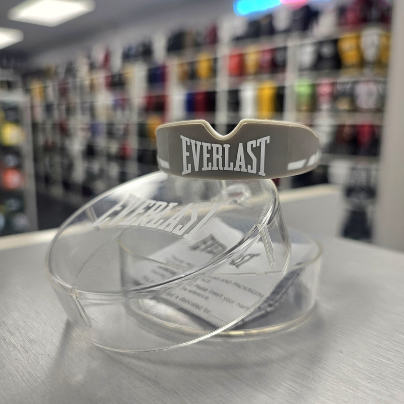 Clear boxing mouthguard with 'Everlast' branding on a metallic surface, blurred sports equipment store background.
