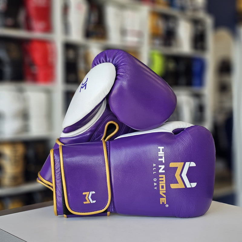 Purple boxing gloves with white and yellow accents, stacked on a white surface with a blurred gym background. The gloves feature the HIT N MOVE brand logo.