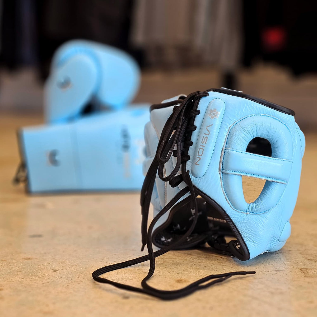 Light blue boxing headgear with black laces in the foreground, with matching boxing gloves blurred in the background on a beige floor.