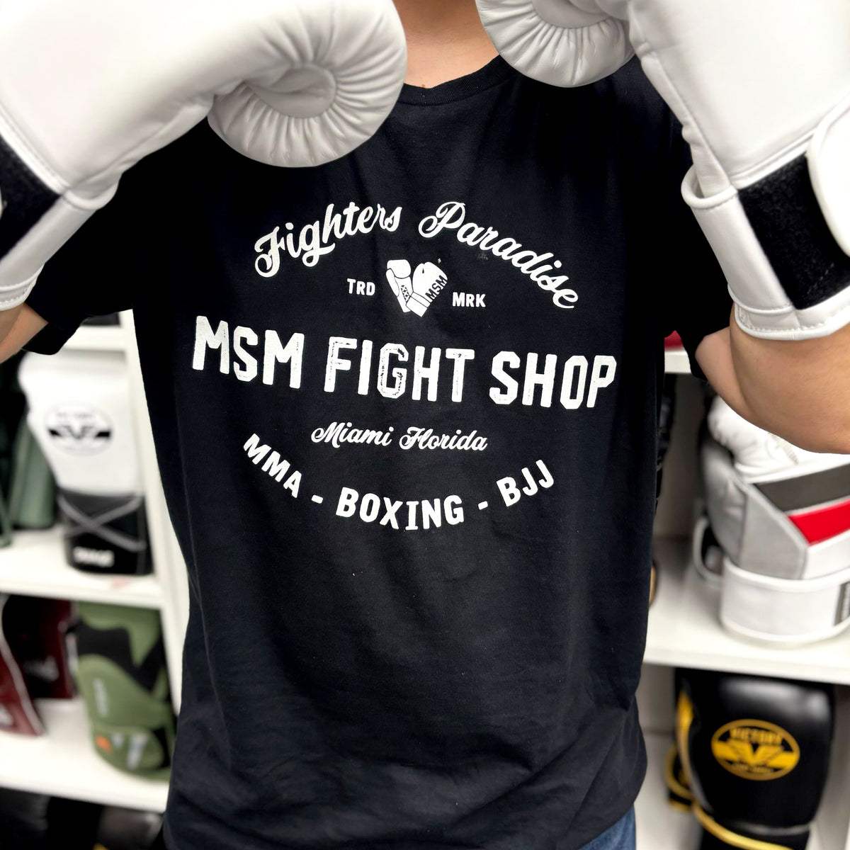Person wearing a black t-shirt with 'MSM Fight Shop' text, white boxing gloves, and gym equipment in the background.