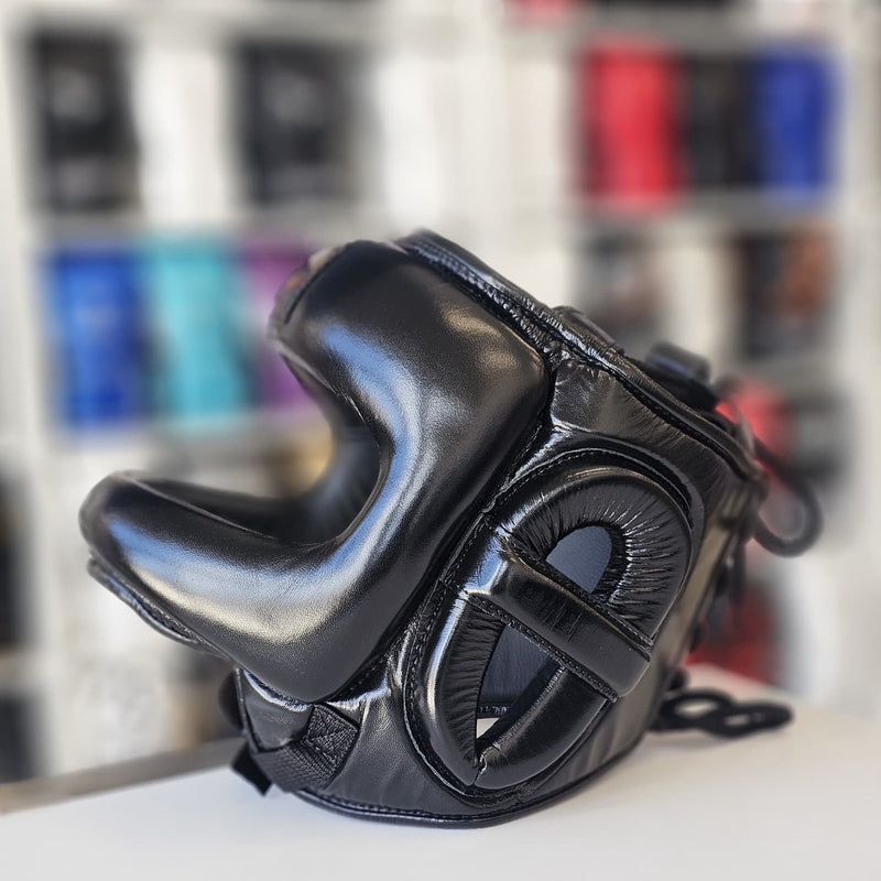 Black leather boxing headgear with padded cheek guards and a chin strap, displayed on a white surface with a blurred background of shelves.