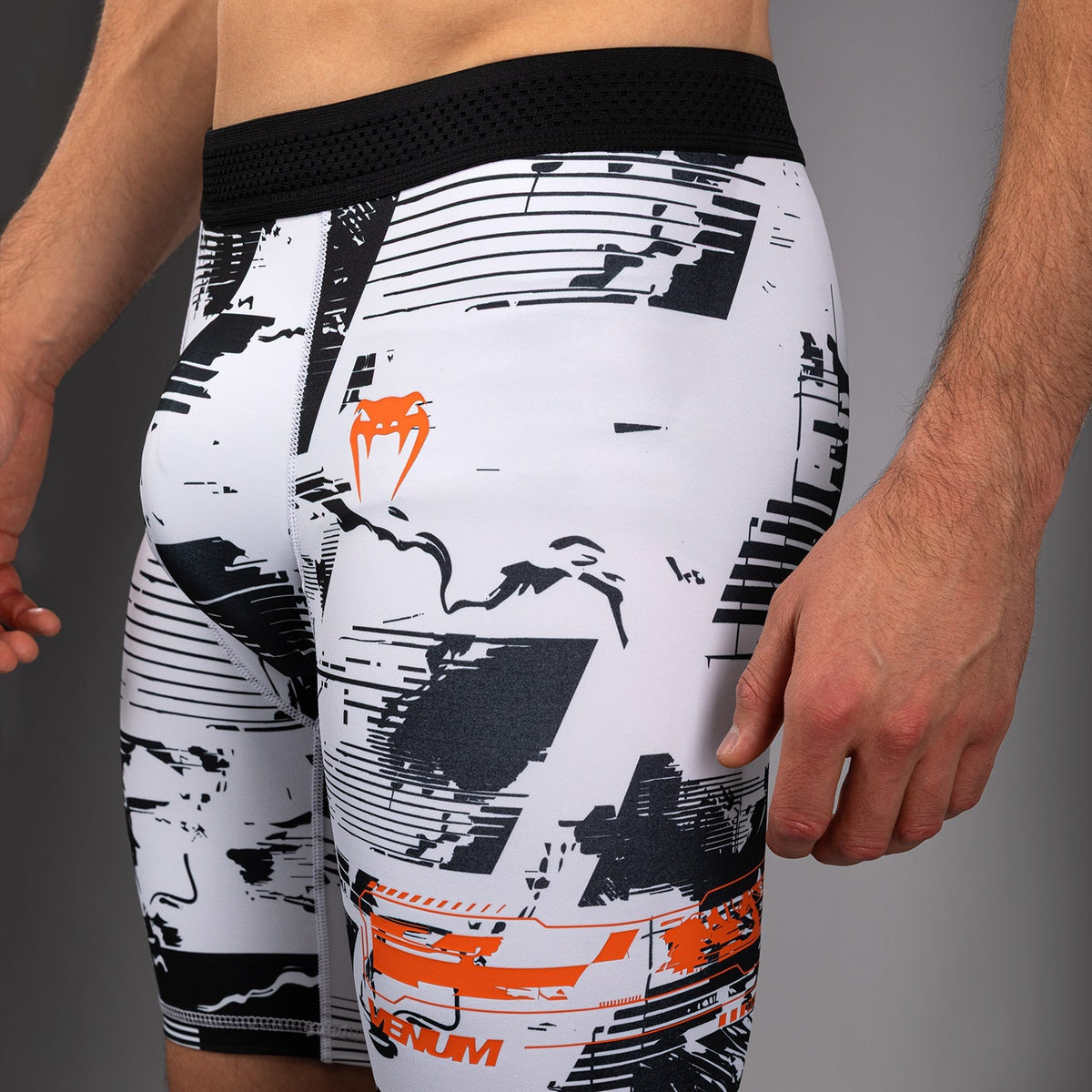 White athletic compression shorts with a black abstract graphic print and orange logo details, shown on a model from the waist down against a gray studio background.