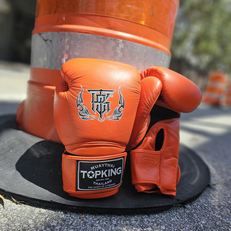 Orange boxing gloves with a brand logo on a black mat, with traffic cones in the background.