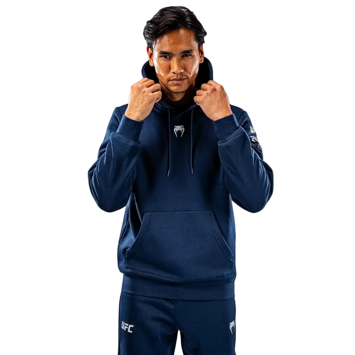 A man wearing a navy blue Venum Fight Gear hoodie and matching sweatpants, with the UFC logo on the sweatpants, posing against a light gray studio background.
