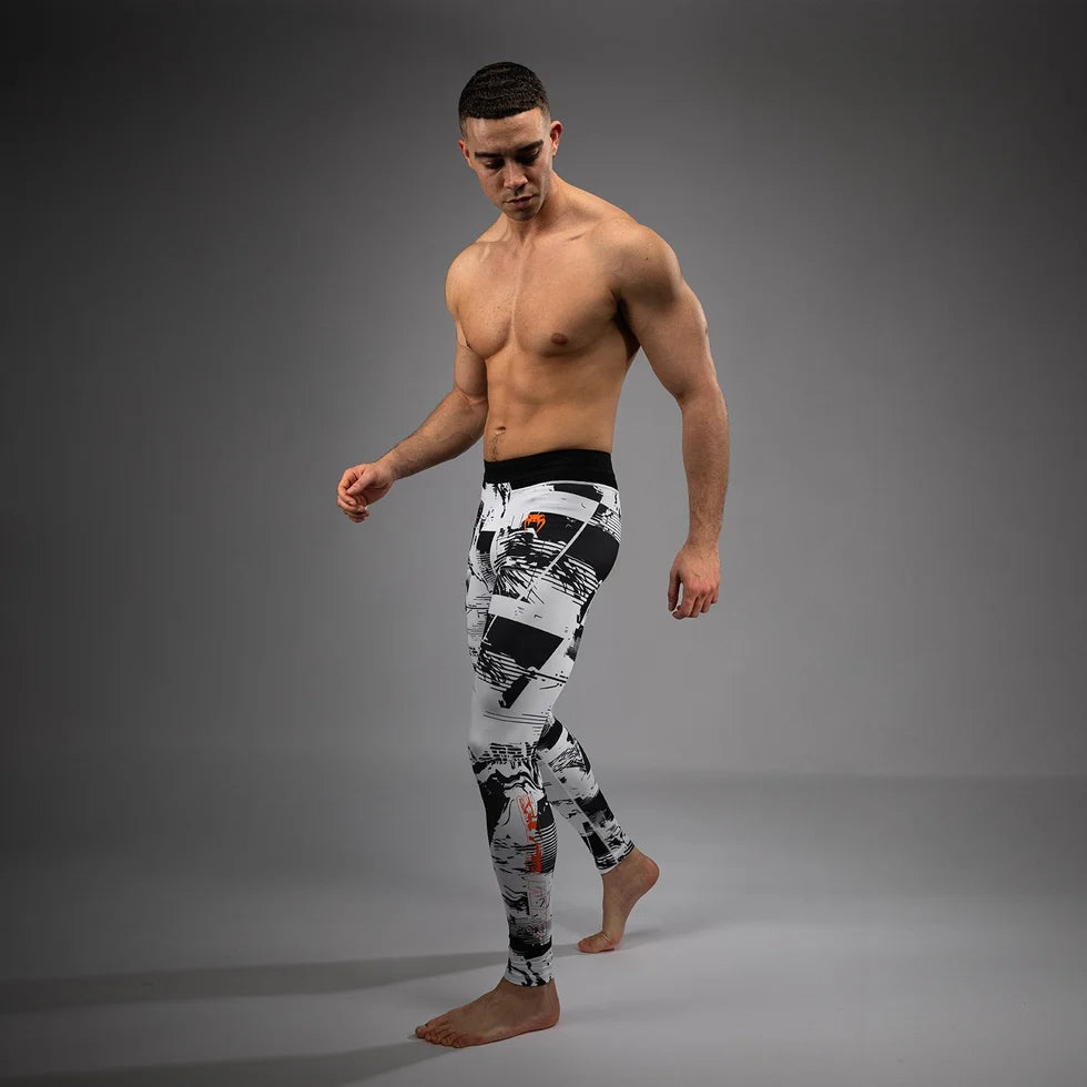 A shirtless male model walking barefoot in black, white, and gray patterned athletic leggings against a dark gray studio background.