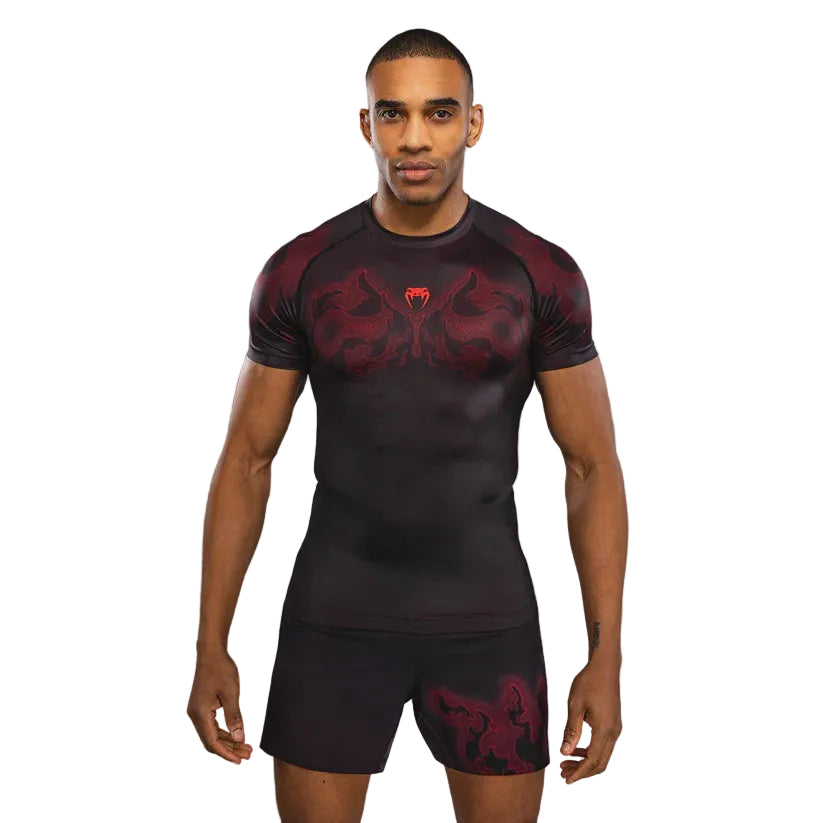 A man modeling a dark gray and red short-sleeve compression shirt with a red snake graphic and a red snake head logo on the chest, paired with matching dark shorts, against a dark gray gradient background.