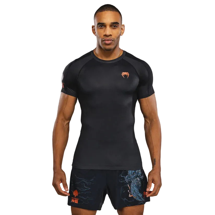 A man modeling a black short-sleeve athletic compression shirt with a small orange logo on the left chest, paired with dark patterned shorts, against a dark gray studio background.