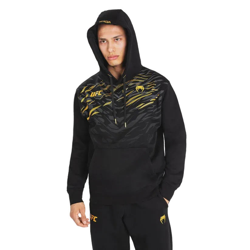 A man wearing a black UFC-branded hooded sweatshirt with gold graphic details and matching black sweatpants, photographed against a dark gray studio background.