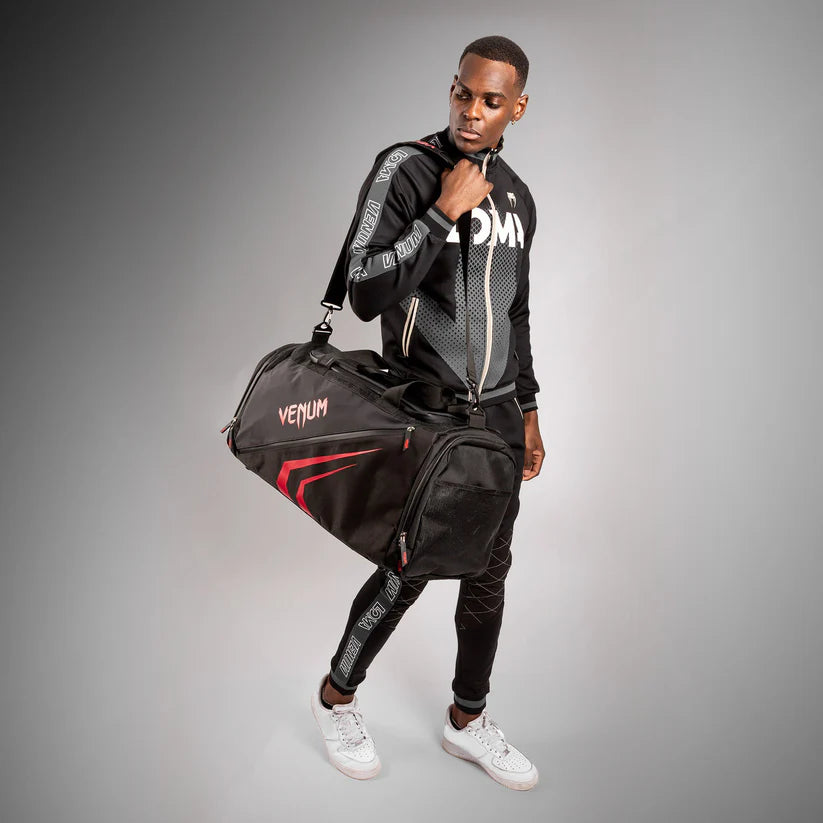 Person wearing a black tracksuit with Venum branding, holding a matching duffel bag on a gray background.