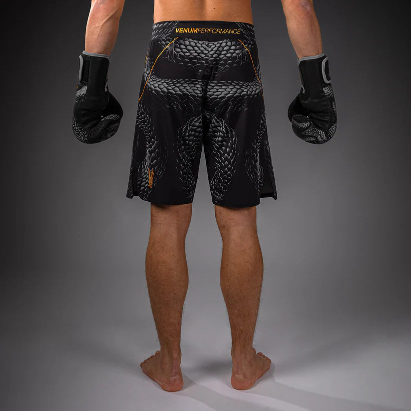 Rear view of a person wearing black Venum fight shorts with a gray snake print and black boxing gloves against a dark gray studio background.