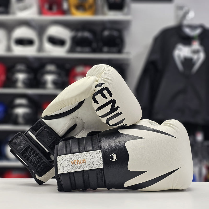 Pair of black and white boxing gloves with 'Venum' branding on a blurred background of sports equipment.