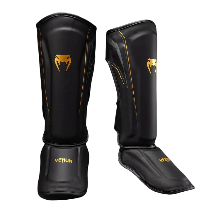 A pair of black Venum shin guards with gold logos and trim, displayed on a clean light gray background.