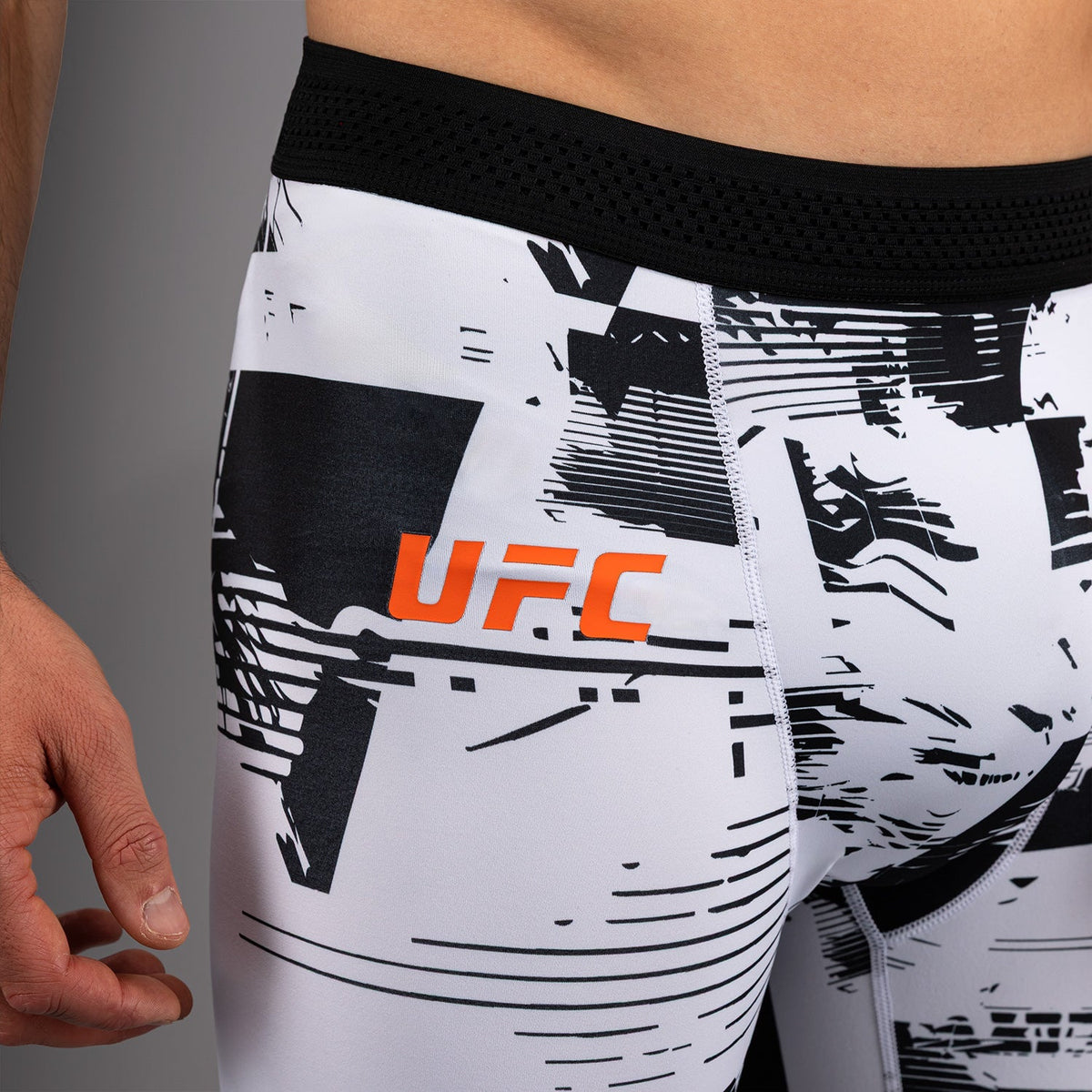 Close-up of a person wearing white UFC athletic compression shorts with a black abstract graphic pattern and a black waistband. The orange UFC logo is printed on the front.
