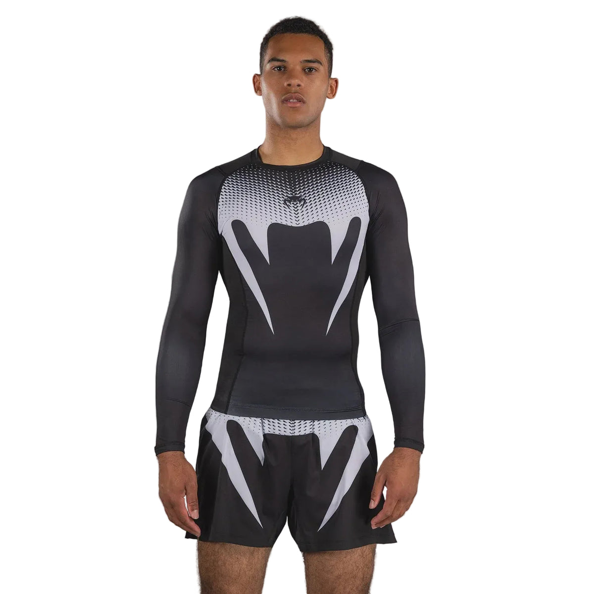 A man modeling a black and white long-sleeve compression rash guard and matching shorts with a Venum logo, set against a solid gray studio background.