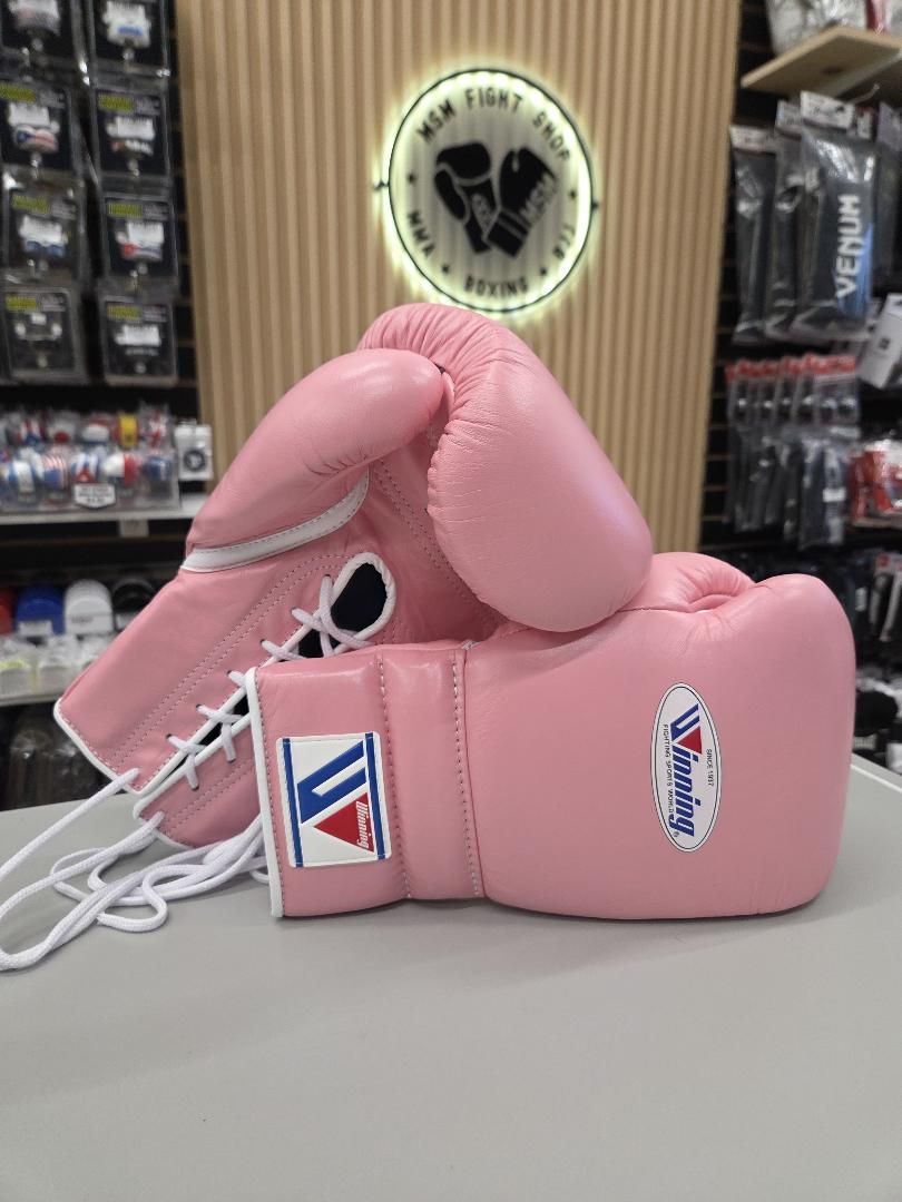 A pair of pink boxing gloves with white laces and visible logos, displayed on a counter inside a retail store with shelves and a sign in the background.