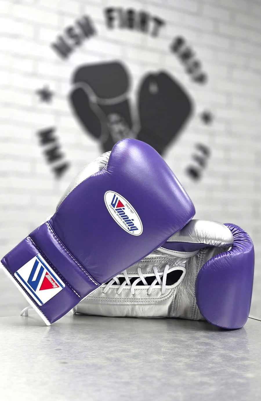 Purple boxing gloves with a brand logo on a gray surface, featuring a blurred background with a logo.