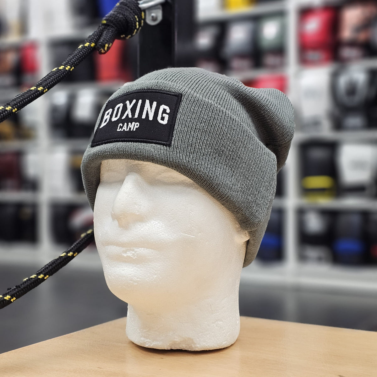 Gray beanie with 'Boxing Camp' patch on a mannequin head against a blurred background of boxing equipment.
